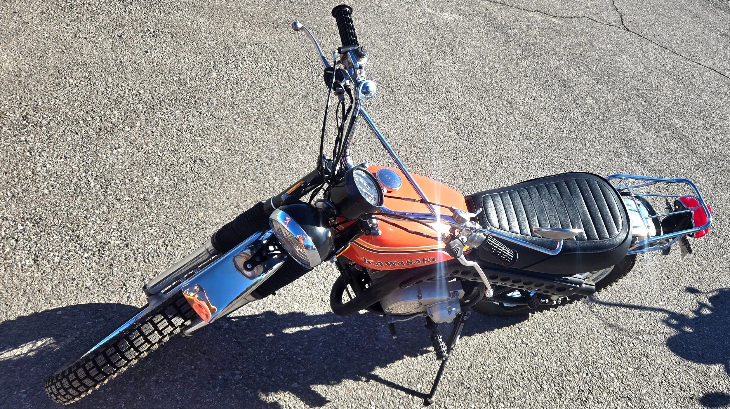 Vintage Kawasaki motorcycle parked on gravel ground, front view, black seat, chrome details, orange fuel tank, black tires.