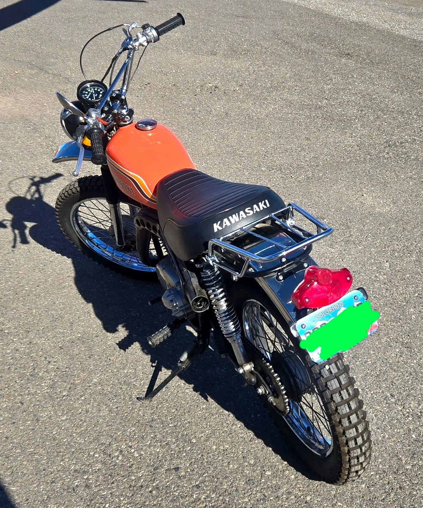 A vintage Kawasaki dirt bike with an orange fuel tank, black seat, and chrome accents, parked on pavement with a Colorado license plate.