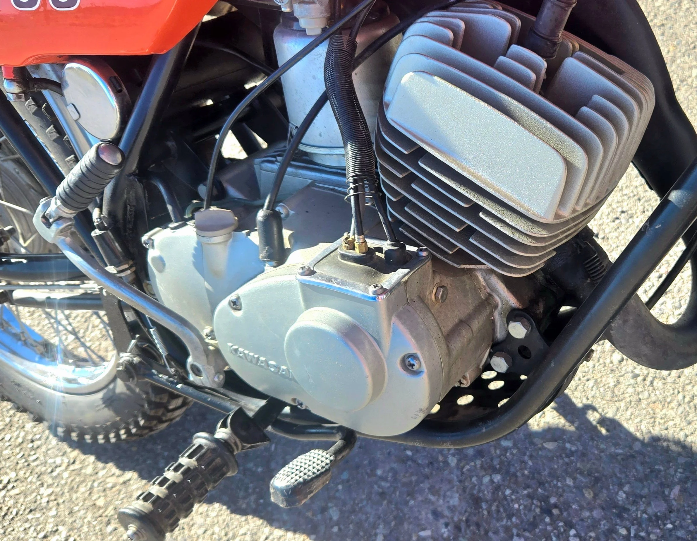 Close-up of a motorcycle engine with visible fins, a kickstart lever, footrest, and logo on the side labeled 'Kawasaki,' on a gravel surface.