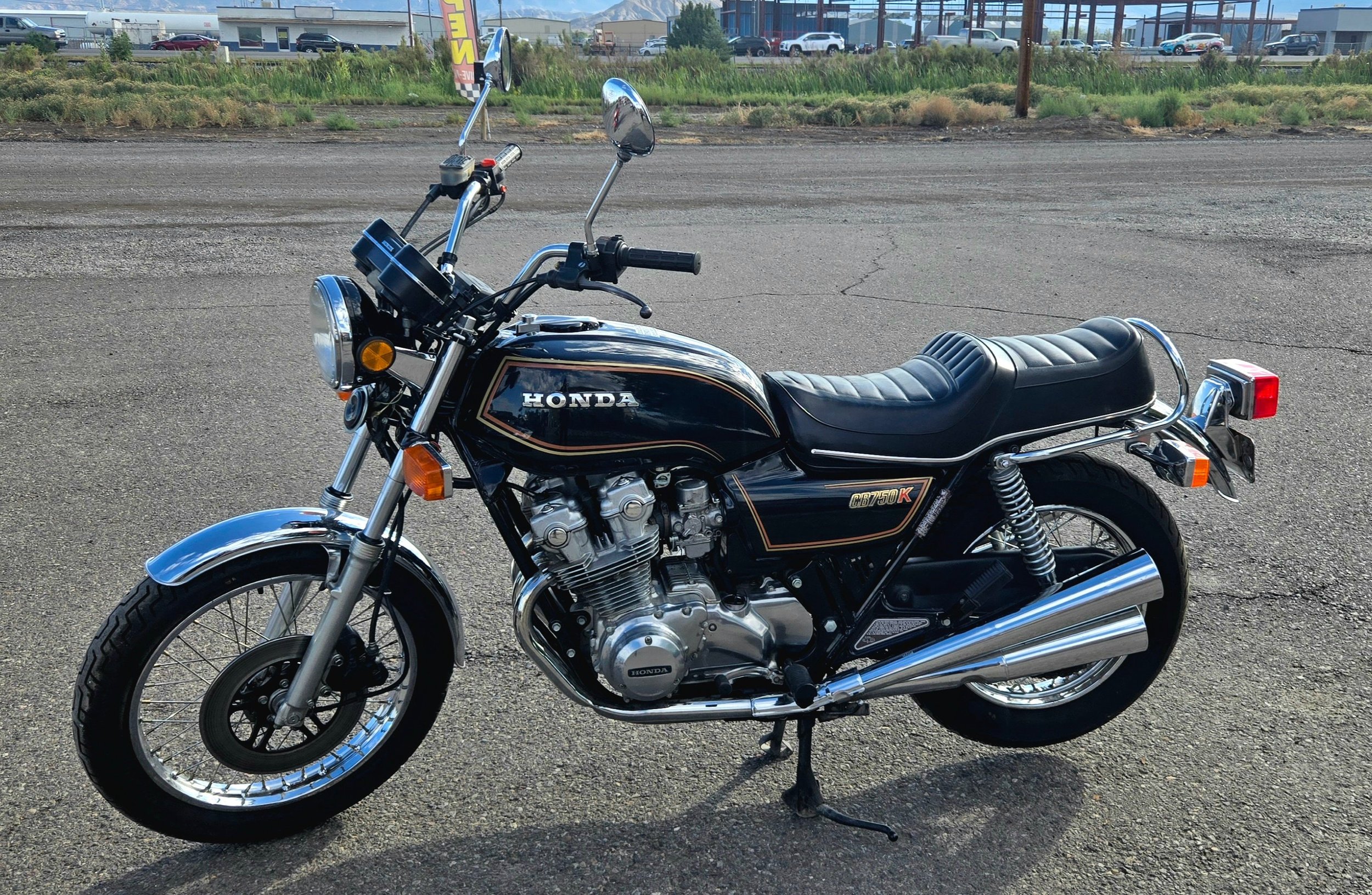 A vintage black Honda motorcycle with chrome details parked on a gravel lot, with a roadside commercial area and mountains in the background.