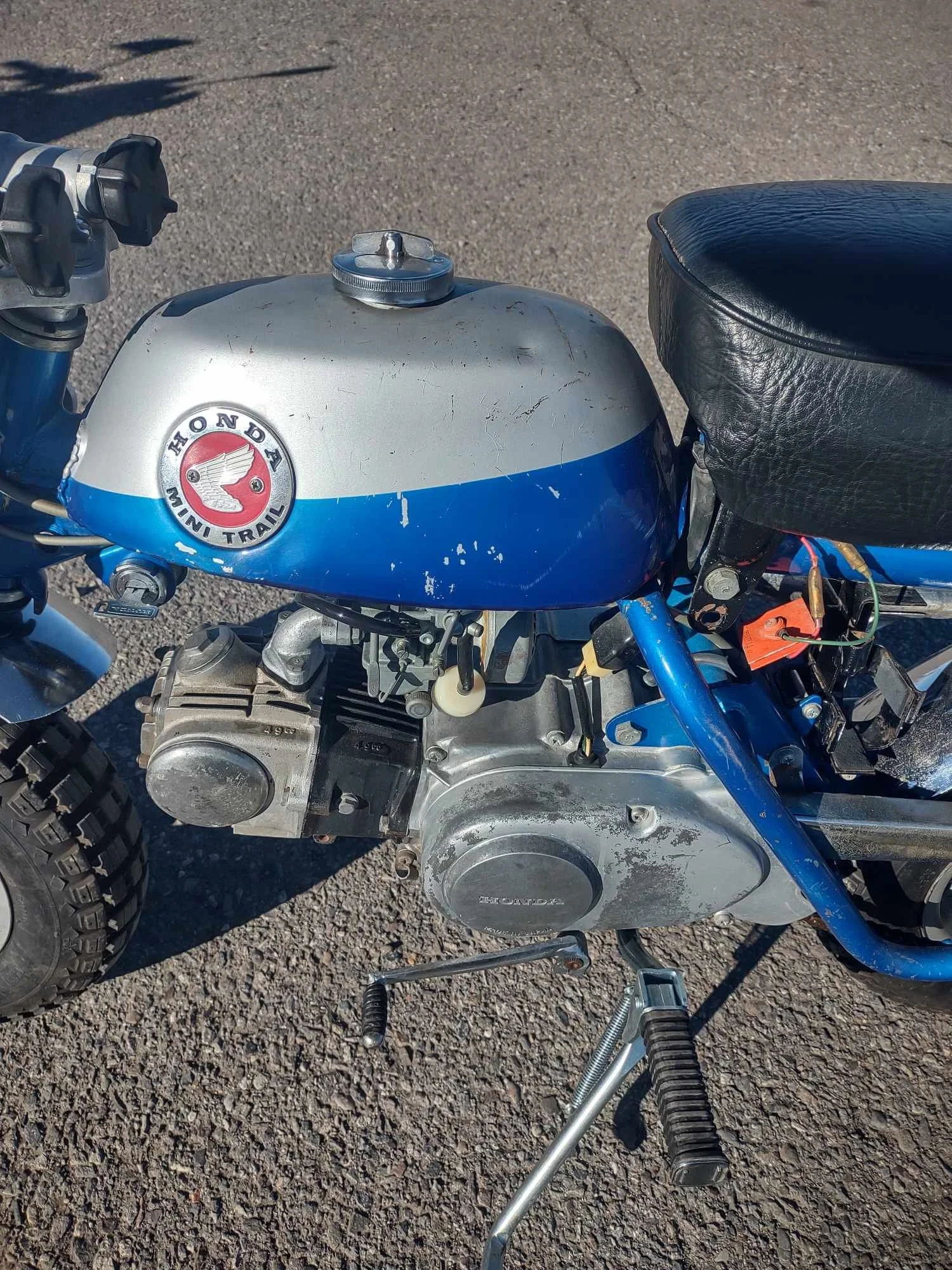 Close-up of a vintage Honda mini trail motorcycle with a silver and blue fuel tank, black seat, and engine, parked on a gravel surface.