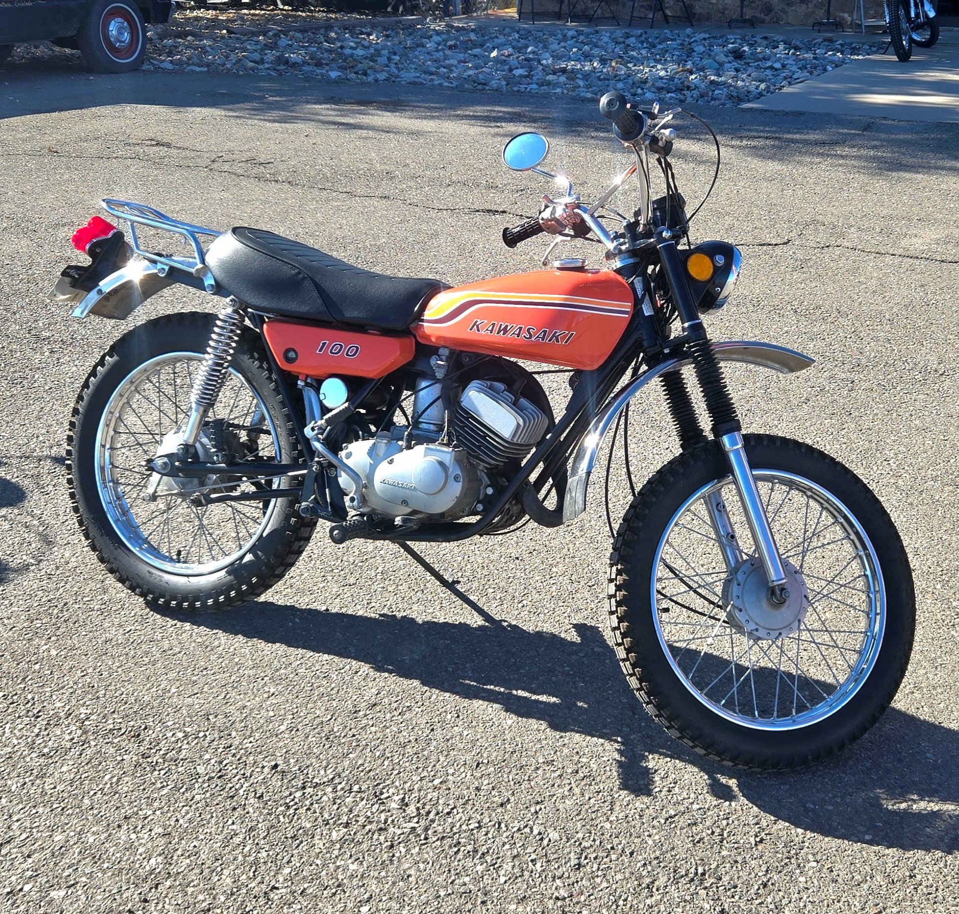 A vintage Kawasaki motorcycle with an orange gas tank, black seat, and knobby tires parked on a gravel surface.
