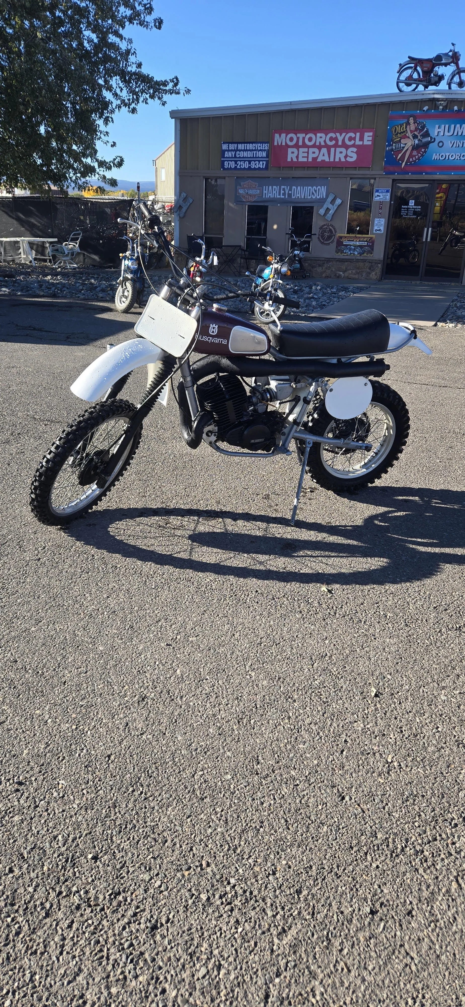 Vintage Husqvarna dirt bike parked on pavement in front of motorcycle repair shop with Harley-Davidson signage.