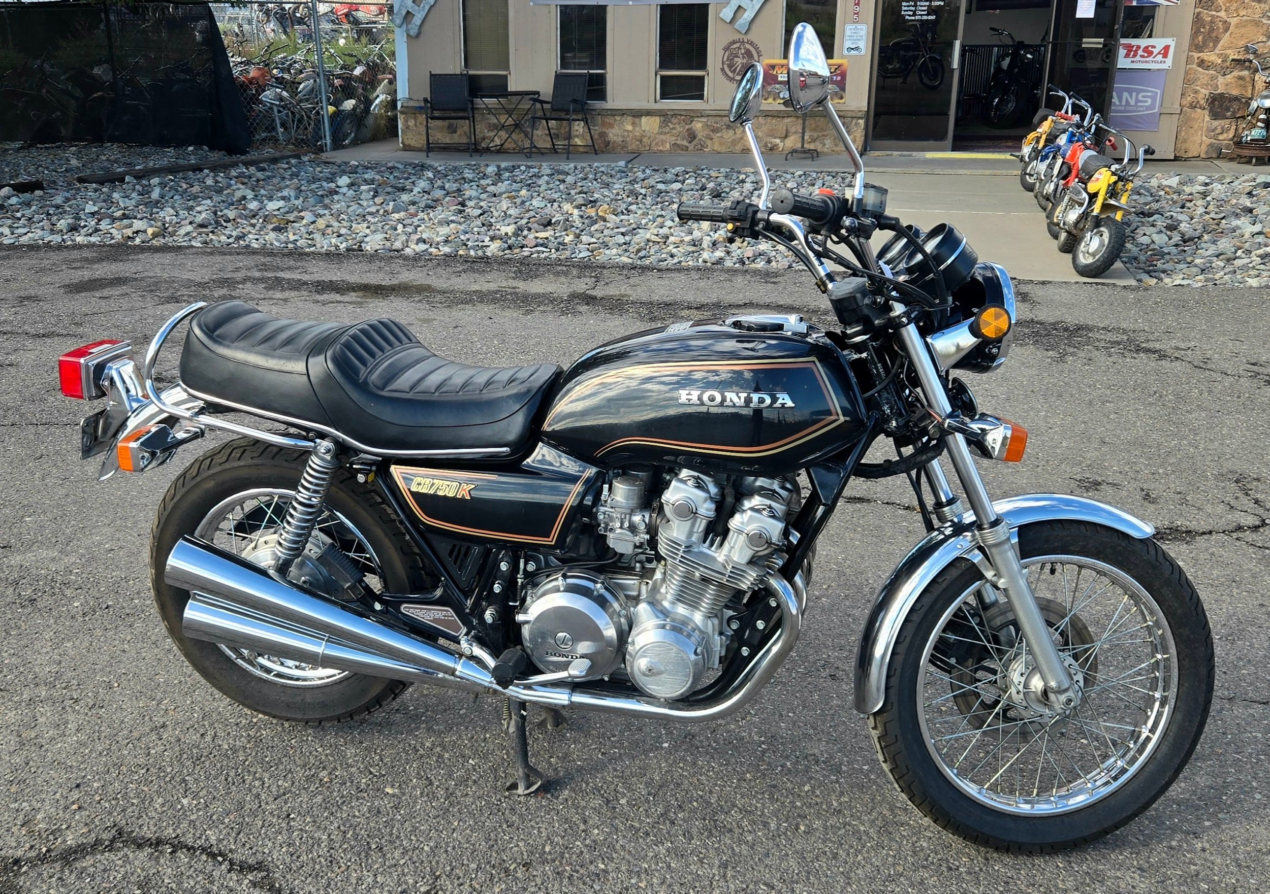 A black vintage Honda motorcycle parked on a street in front of a motorcycle shop. The motorcycle has chrome accents, a black leather seat, and a chrome exhaust, with in the background a small building, a few chairs, and a row of small motorbikes.