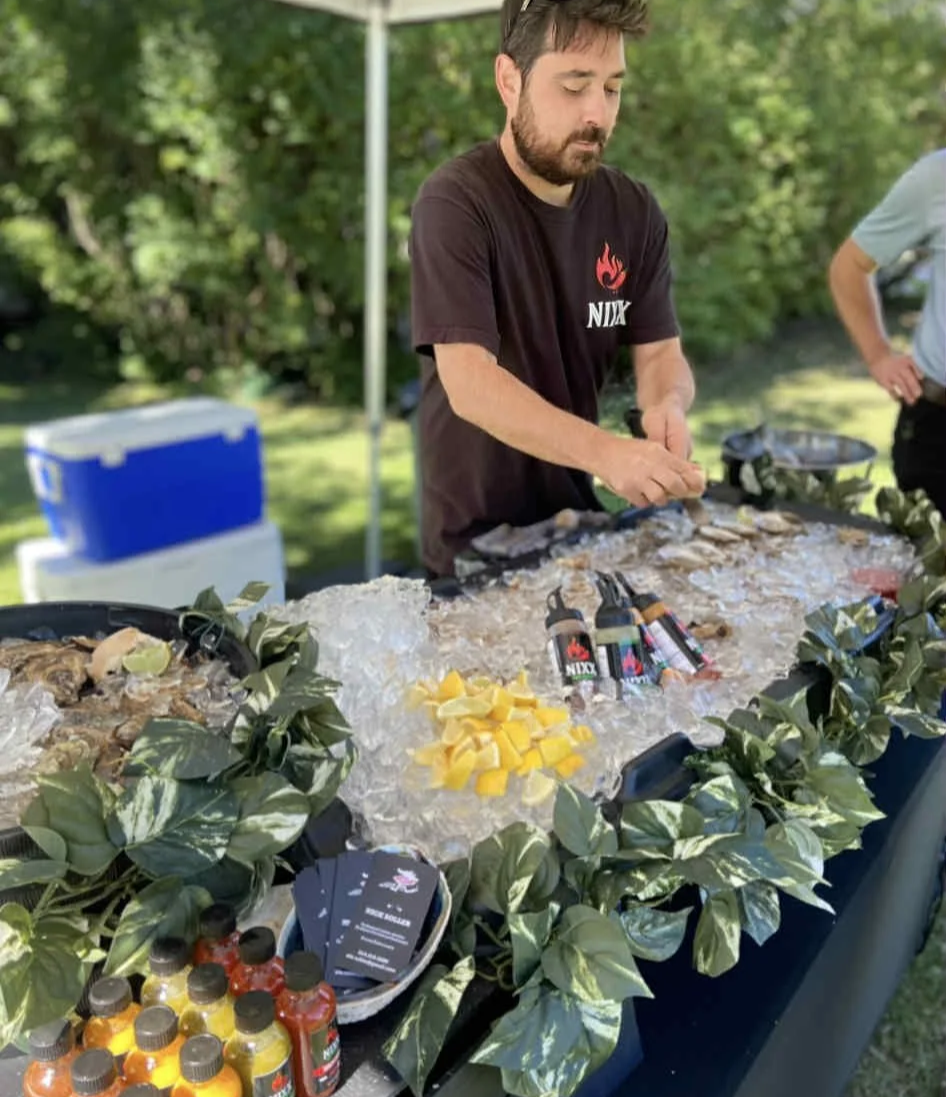 A man standing at a table outside, preparing food with ice and bottled drinks, with another person partially visible to the right; the table is decorated with green foliage.