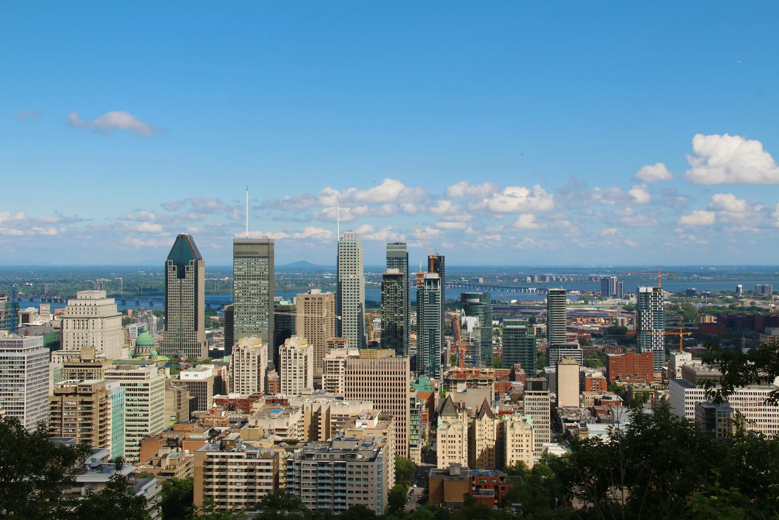 Aerial view of a city skyline with tall skyscrapers, some under construction, set against a bright blue sky with scattered clouds, and a river in the background.