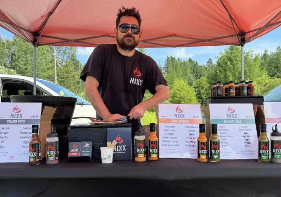 A man wearing sunglasses and a black NIXX T-shirt stands behind a food stall under a pink canopy, with bottles of NIXX sauces and menus displayed on the table. Trees and cars are visible in the background.
