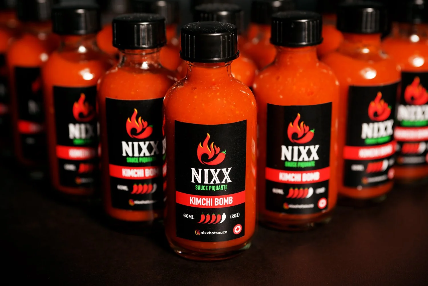 Multiple bottles of NIXX Kimchi Bomb hot sauce arranged on a dark surface, featuring black labels with a flame logo and red, green, and white text.