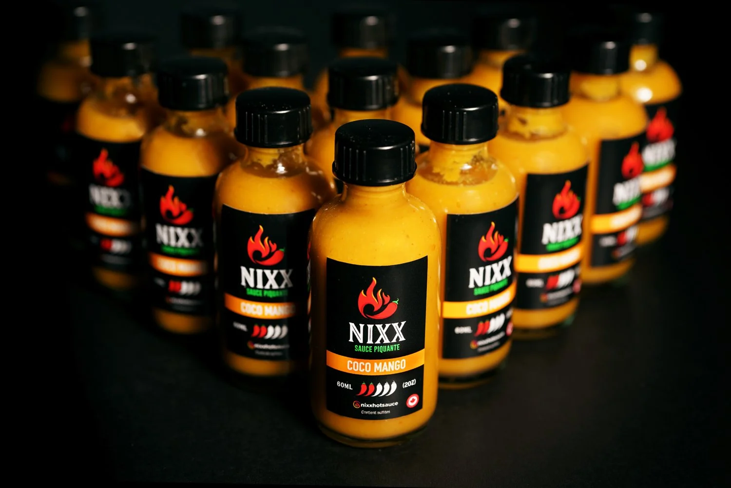 A row of bottles of NIXX hot sauce with a label indicating 'Coco Mango' flavor, black caps, and black labels with red chili flame logo on black background.