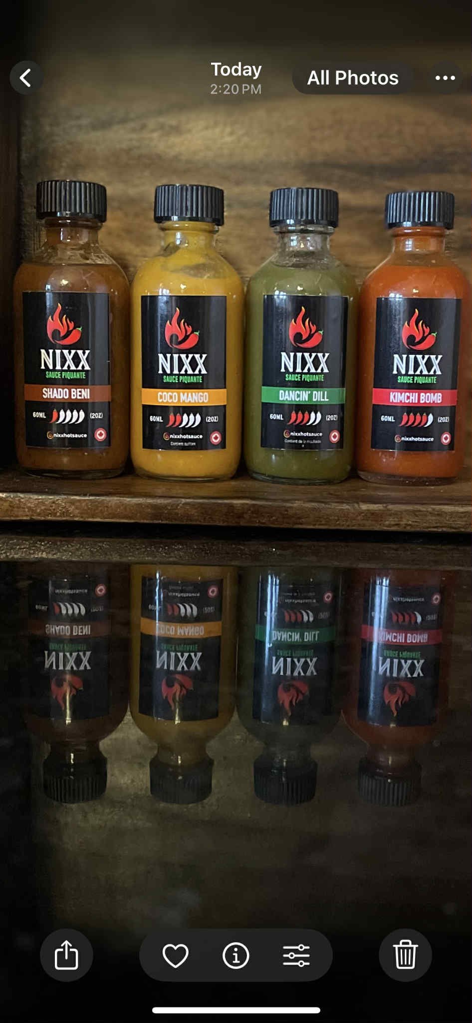 Four bottles of hot sauce labeled with different flavors: Shado Beni, Coco Mango, Dancin' Dill, and Kimchi Bomb, placed on a wooden shelf.