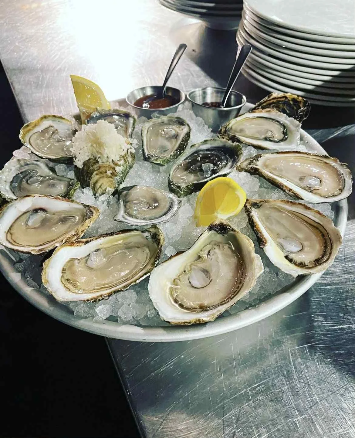 A large white plate with ice and fresh oysters, lemon wedges, and two small cups of cocktail sauce on a stainless steel table.