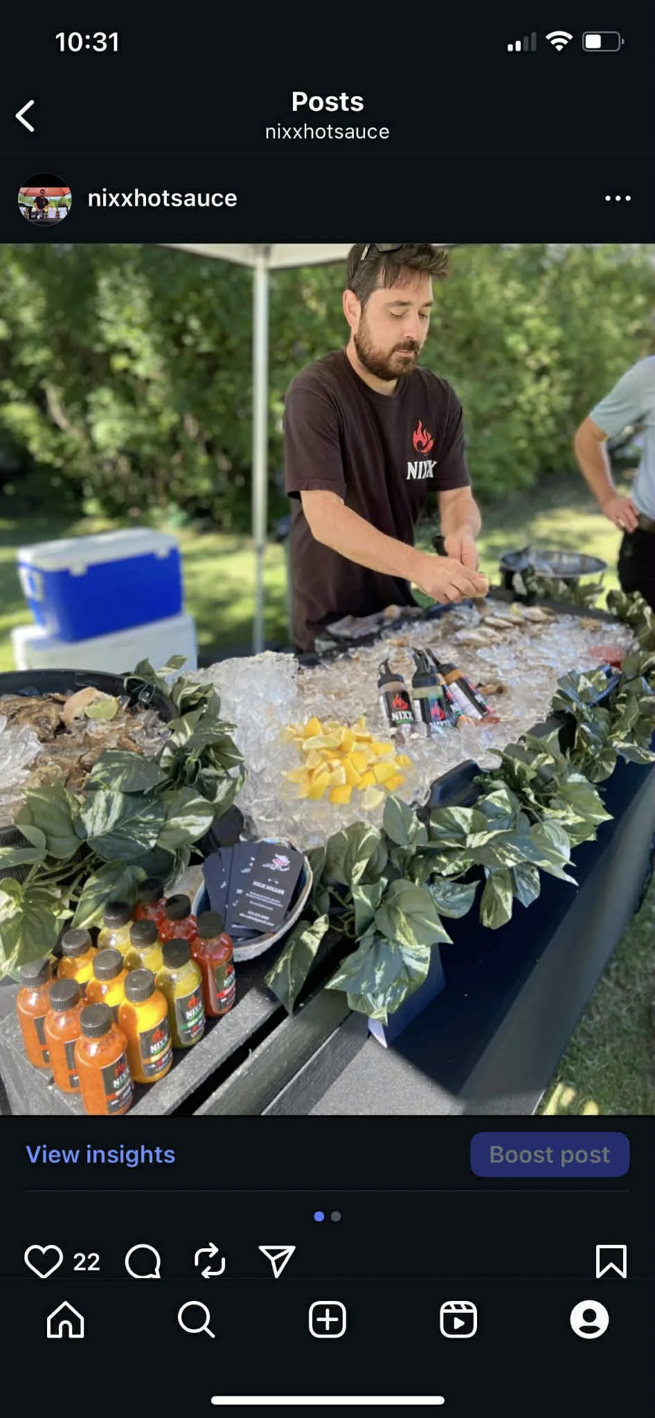 A man with a beard and dark hair preparing grilled food at an outdoor barbecue stand decorated with green foliage, bottles of hot sauce, and lemon wedges, with cooler and another person in the background.