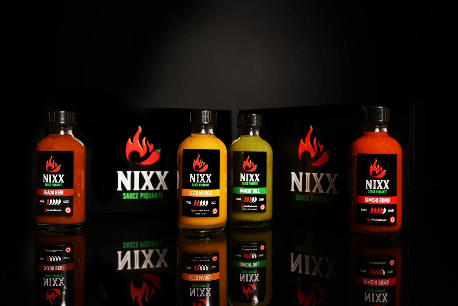 Four bottles of spicy sauce labeled NIXX with different flavors: Shado Beni, Coco Mango, Dancin' Dill, and Kimchi Bomb, standing on a reflective black surface with their labels and reflections visible.
