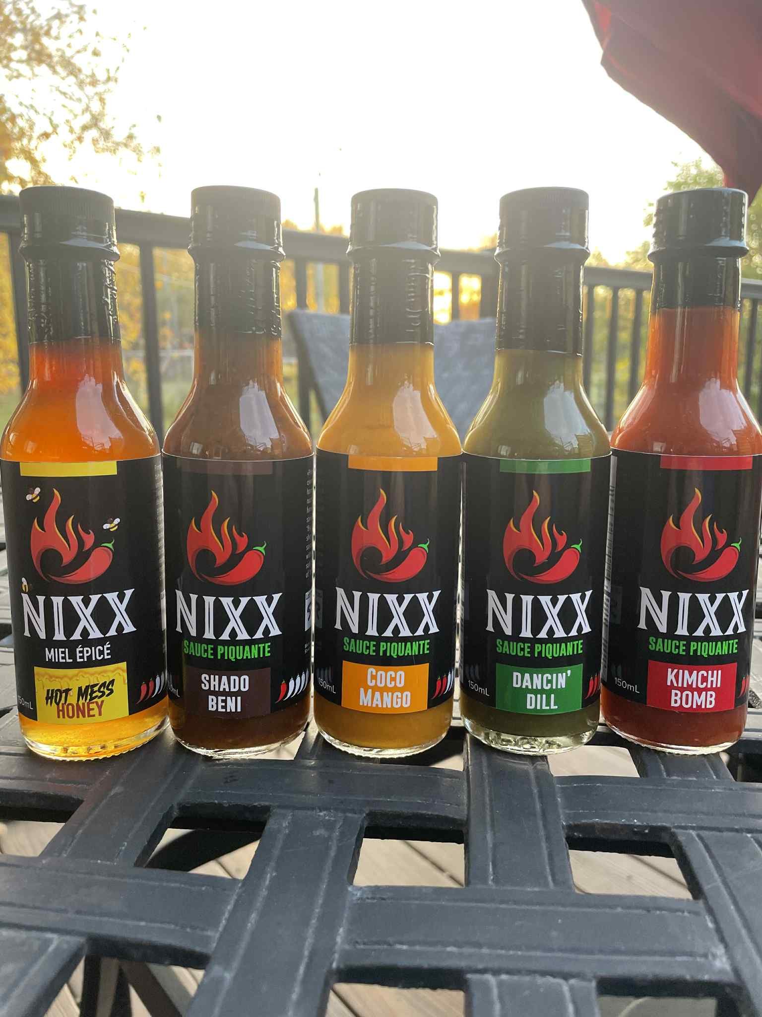 Five bottles of NIXX hot sauce and condiments, labeled 'Hot Mess Honey,' 'Shado Beni,' 'Coco Mango,' 'Dancin Dill,' and 'Kimchi Bomb,' placed side by side on an outdoor table with a railing and trees in the background during sunset.