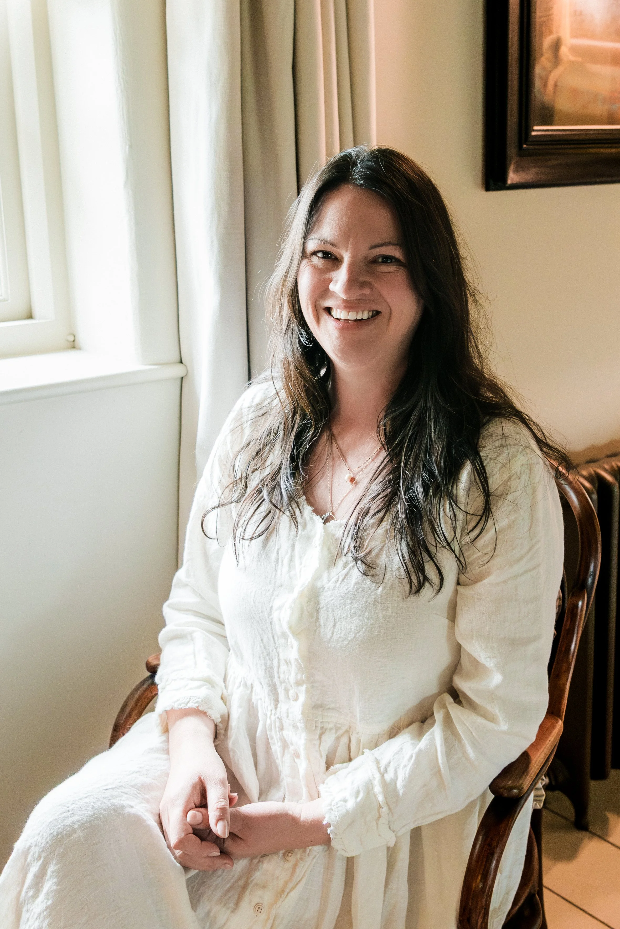 A woman with long, dark wavy hair smiling and sitting on a wooden chair by a window with beige curtains.