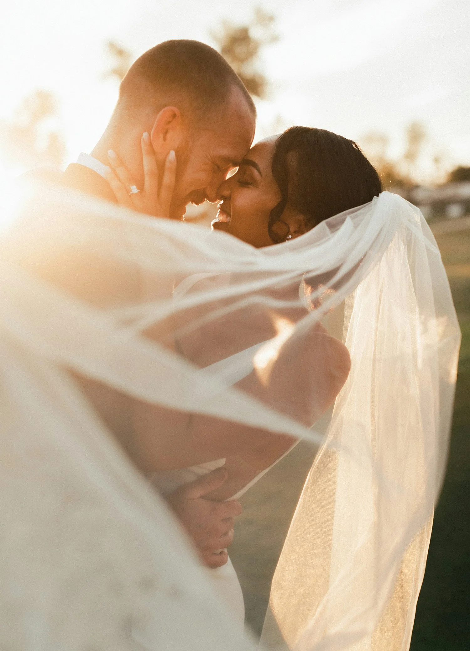 A joyful bride and groom embrace each other outdoors during sunset, wrapped in a veil, sharing a close, happy moment.