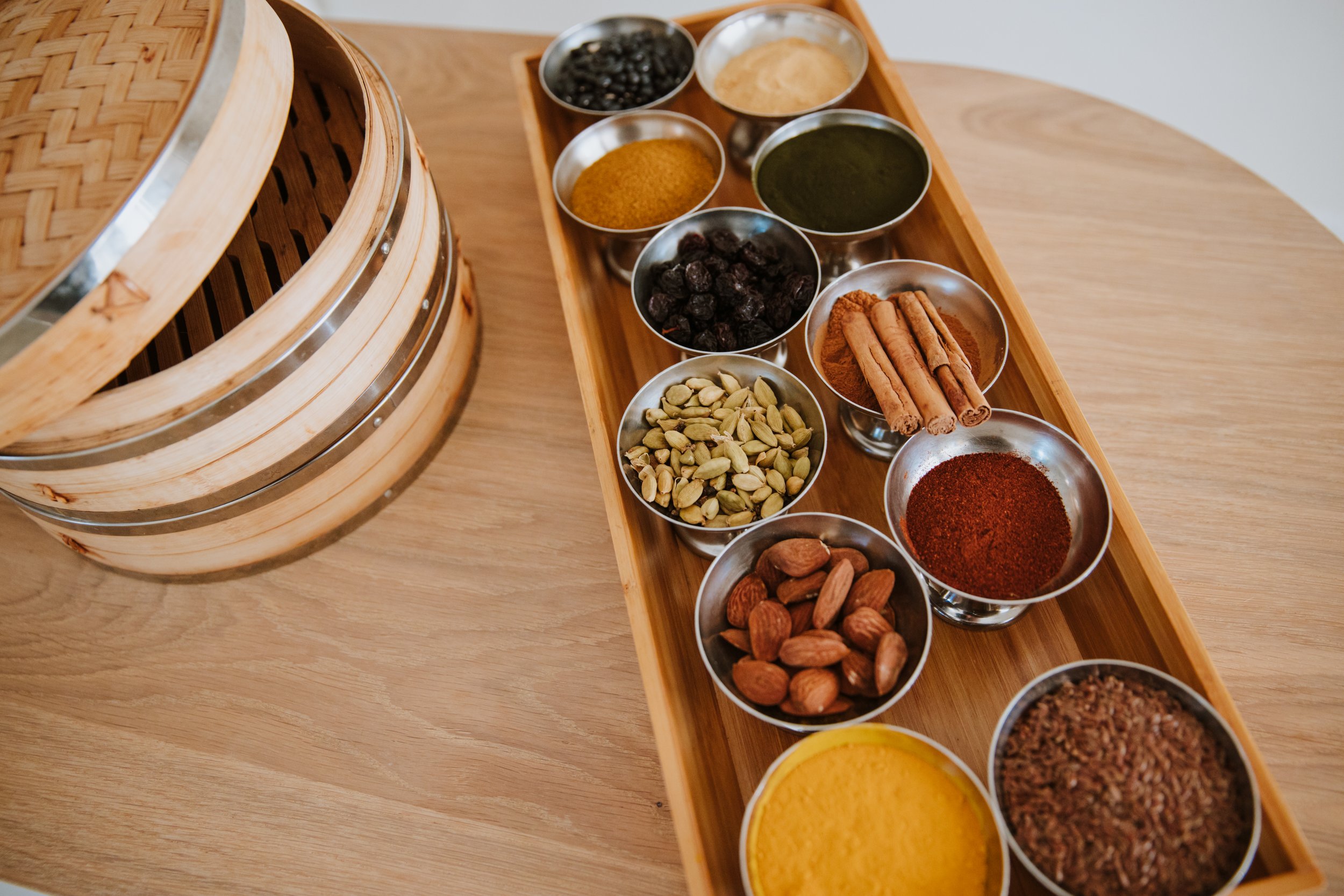 Varie spezie e ingredienti in ciotole di metallo, tra cui cardamomo, pistacchi, mandorle, curcuma, curry, cannella, pepe, sesamo, paprika, zenzero, noce moscata, e altri, con un contenitore di bambù accanto.