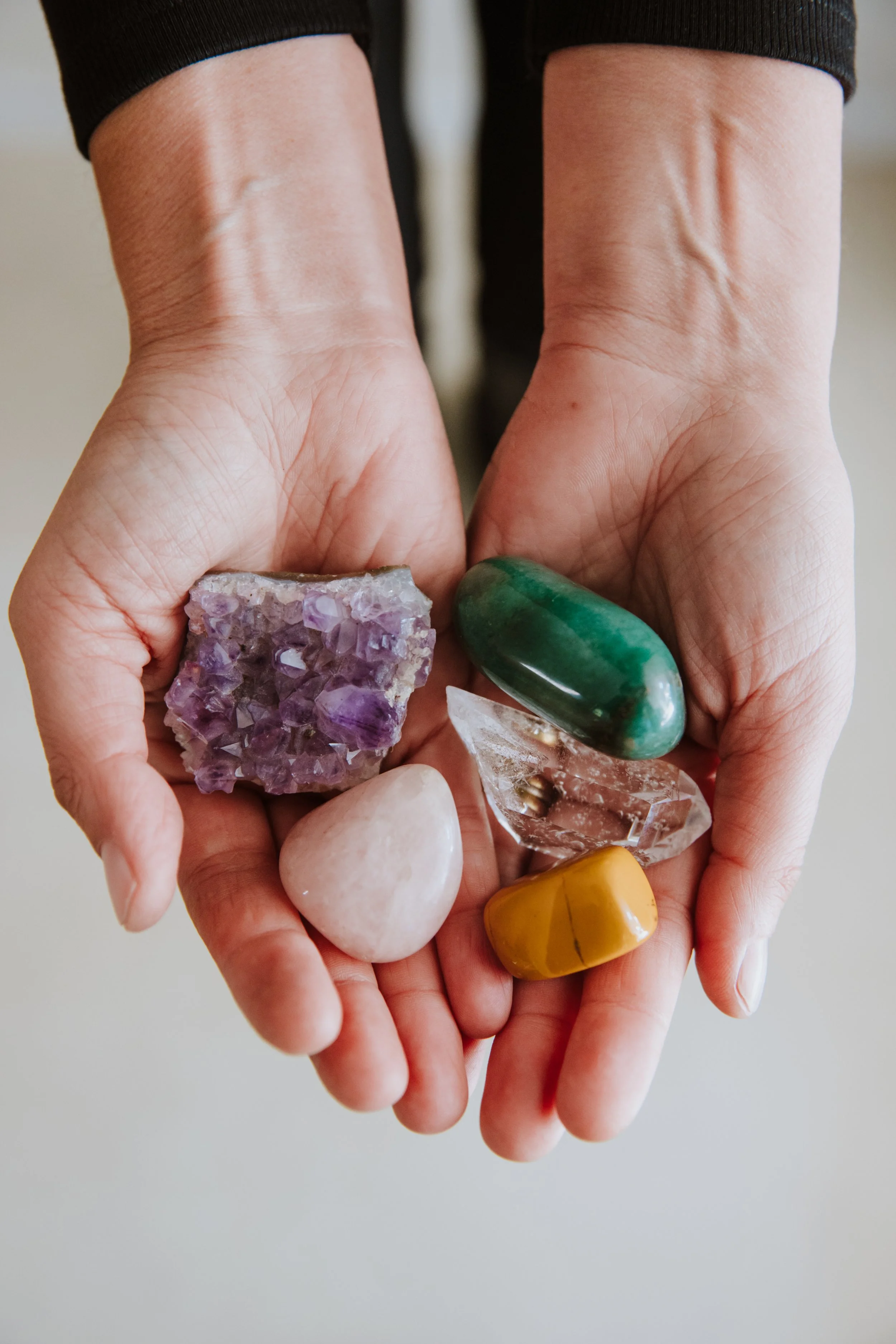 Due mani che tengono diverse pietre e cristalli colorati, tra cui un quarzo rosa, una pietra verde, un ametista e altre.