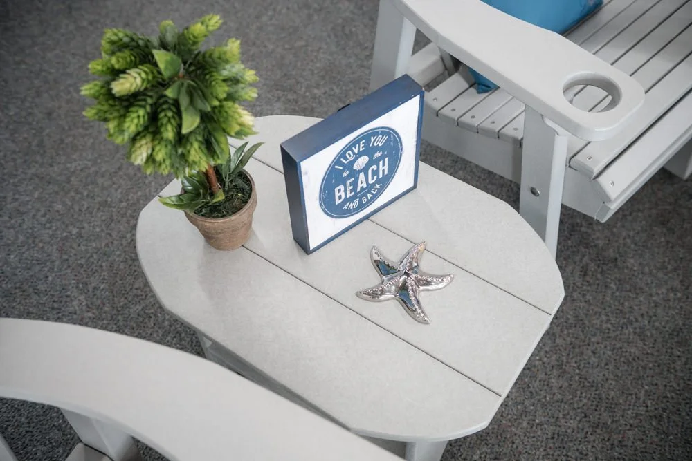 A white outdoor table with a small potted green plant, a framed sign that says 'Love you to the beach and back,' and two starfish ornaments, situated near a white bench on gray carpet.