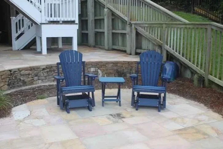 Two blue Adirondack chairs with a small matching table in between, placed on a stone patio in a backyard with a deck and wooden railing in the background.