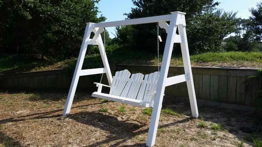White wooden outdoor swing with a backrest shaped like three planks, hanging from a white frame in a backyard with grass and trees.