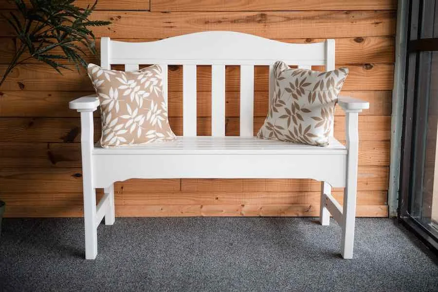 White wooden bench with two beige and white floral pillows, set against a wooden wall and next to a glass door.