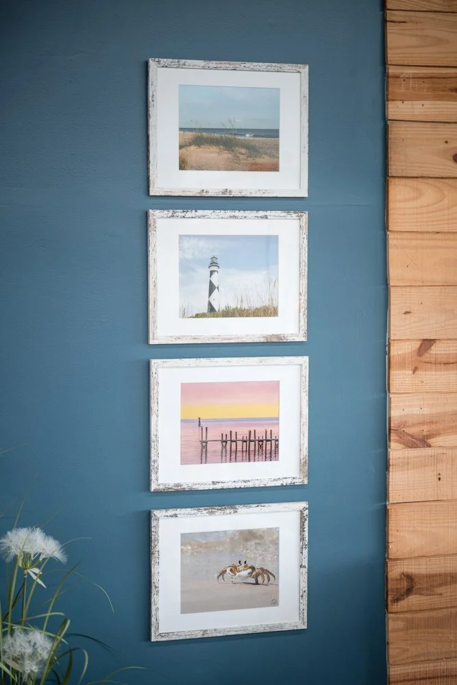 Four framed coastal-themed photographs hung vertically on a blue wall, with a wooden panel on the right side of the wall.