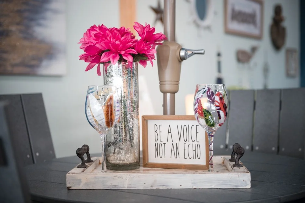 Decorative tray with pink flowers in a silver vase, two wine glasses with colorful leaves and feathers, a framed quote that says "Be a voice, not an echo," and a champagne opener, on a dark table.