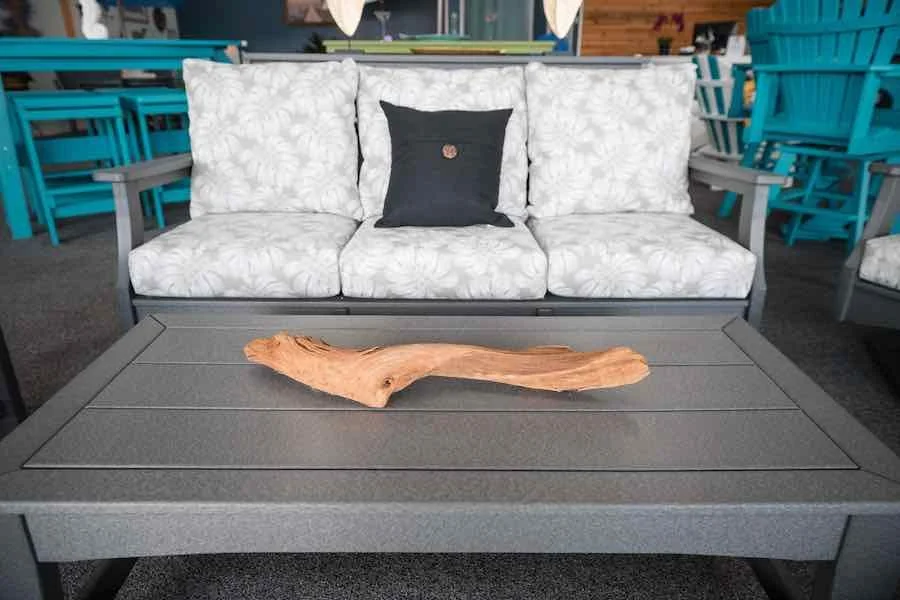Outdoor patio furniture set includes a cushioned sofa with three white floral patterned cushions and a single black cushion, a gray metal coffee table with a wooden decorative piece on top, and surrounding turquoise chairs in a retail or furniture store.