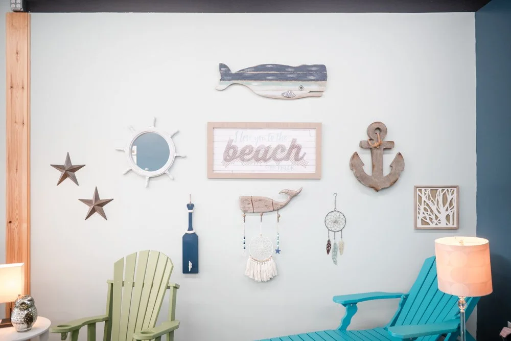 Beach-themed interior room with wall decorations including a wooden fish, an anchor, stars, a mirror, a sign that says 'Hello you to the beach,' a small paddle, a whale figure, a dreamcatcher, and a framed art piece. There are two Adirondack chairs, 