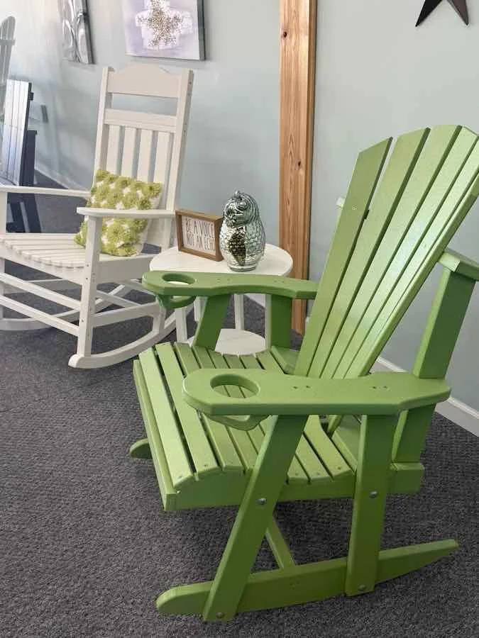 Two Adirondack chairs, one green and one white, placed on a carpeted floor inside a room. The green chair is in the foreground, and the white one is in the background, with a small table between them. The table holds a clear vase with a decorative twig, a framed sign that says "BE KIND, BE HONEST," and a framed picture. A wall with artwork and a black star decoration are visible on the walls.