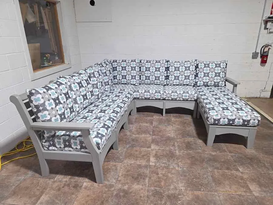 Grey L-shaped outdoor patio bench with floral cushions in a room with white brick wall and tiled floor.