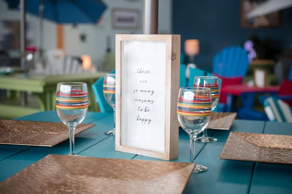 Table setting with four glasses of water, woven placemats, and a framed sign that reads 'There are so many reasons to be happy.' in a colorful, casual restaurant or cafe.