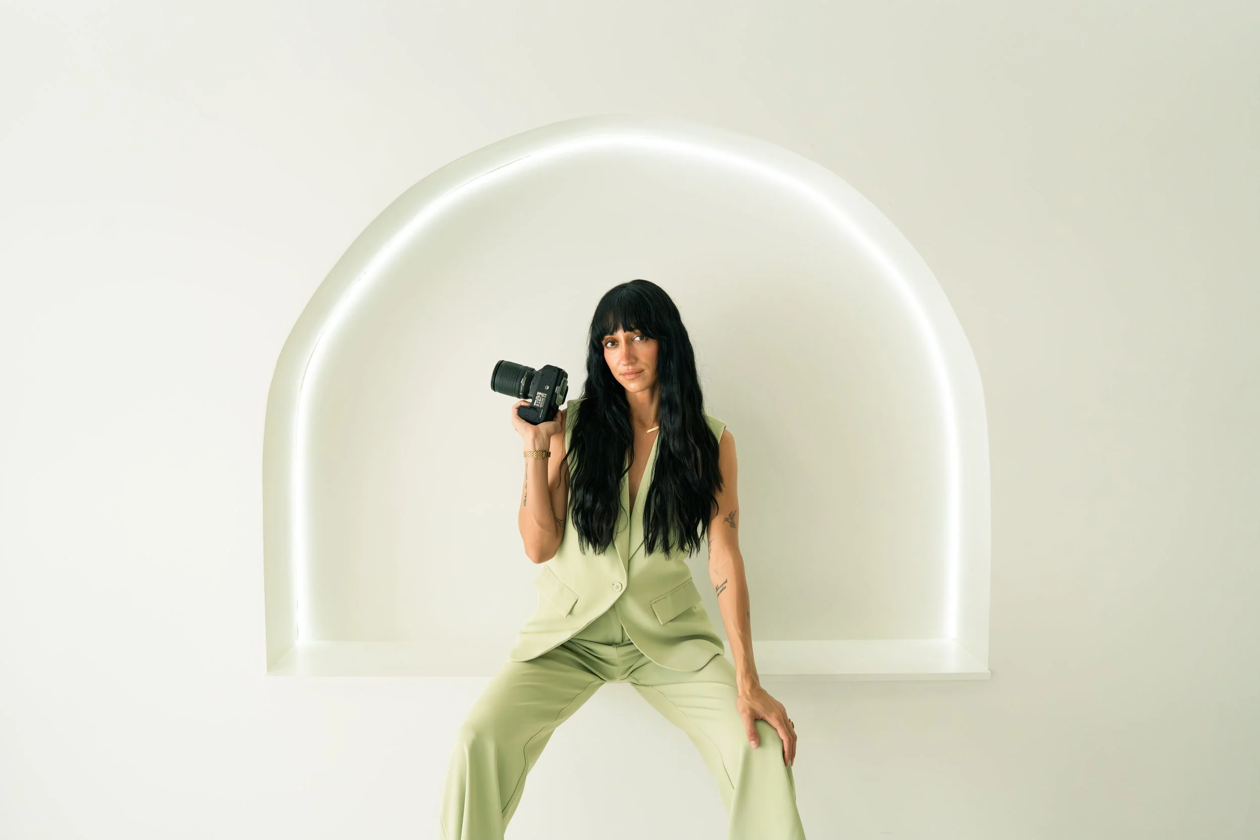 Woman with long black hair holding a camera, dressed in a light green sleeveless suit, sitting on a ledge in front of a white wall with a rounded neon light arch