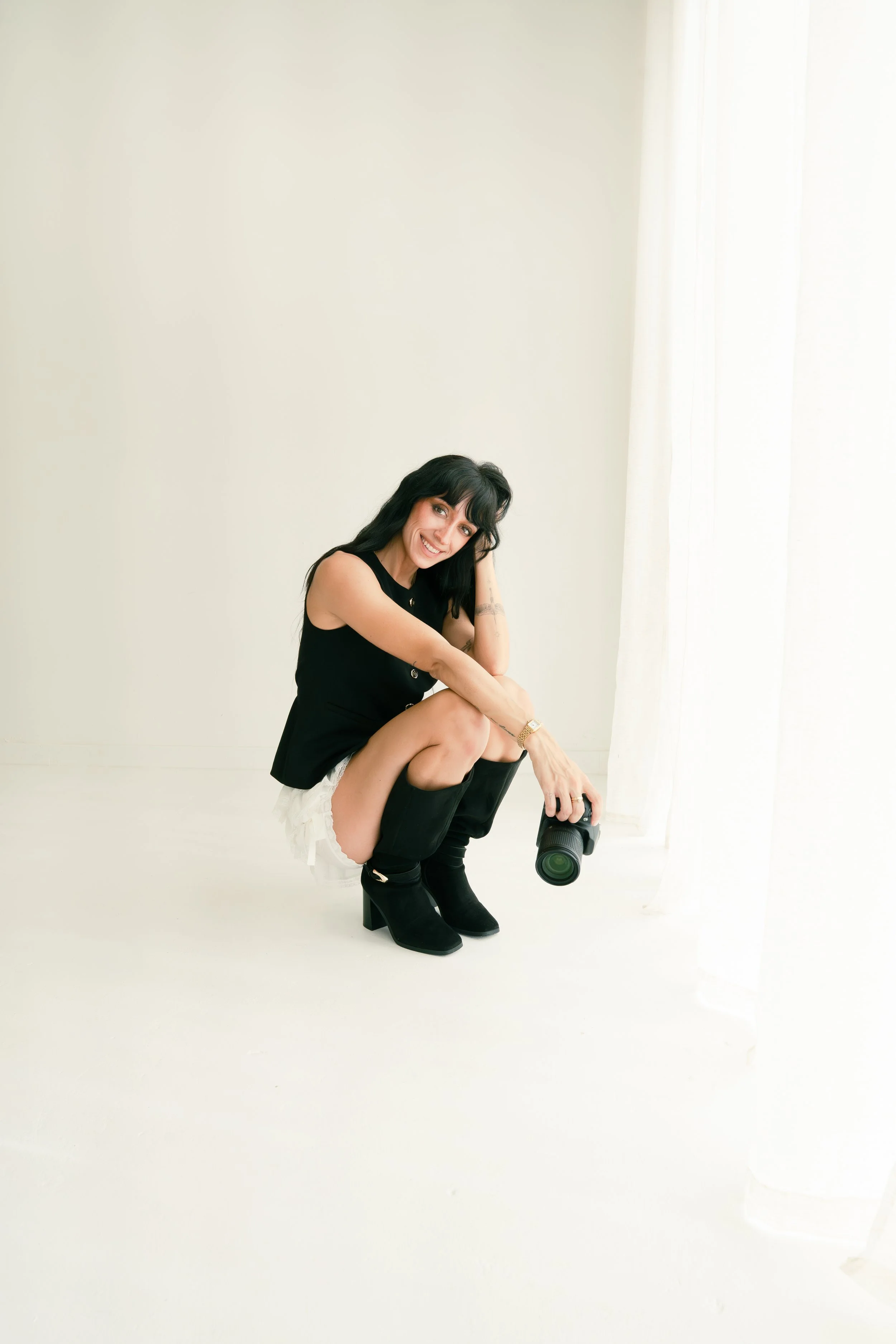 Woman crouching, holding a camera, smiling, with black hair, wearing a black sleeveless top, white shorts, and black heeled boots, in a bright room with white walls and sheer curtains.