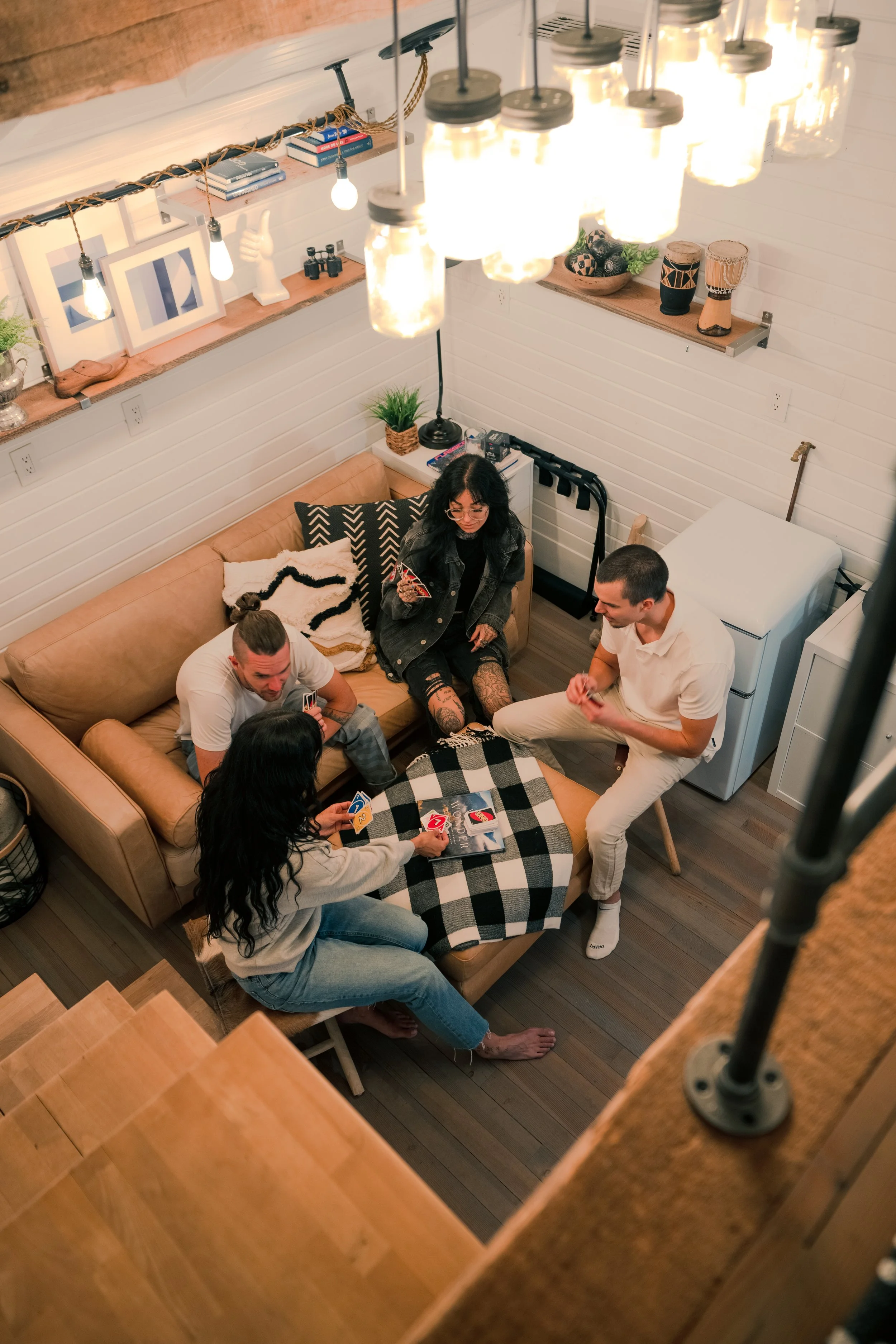Four people playing a card game around a checkered table in a cozy living room with warm lighting and modern decor.