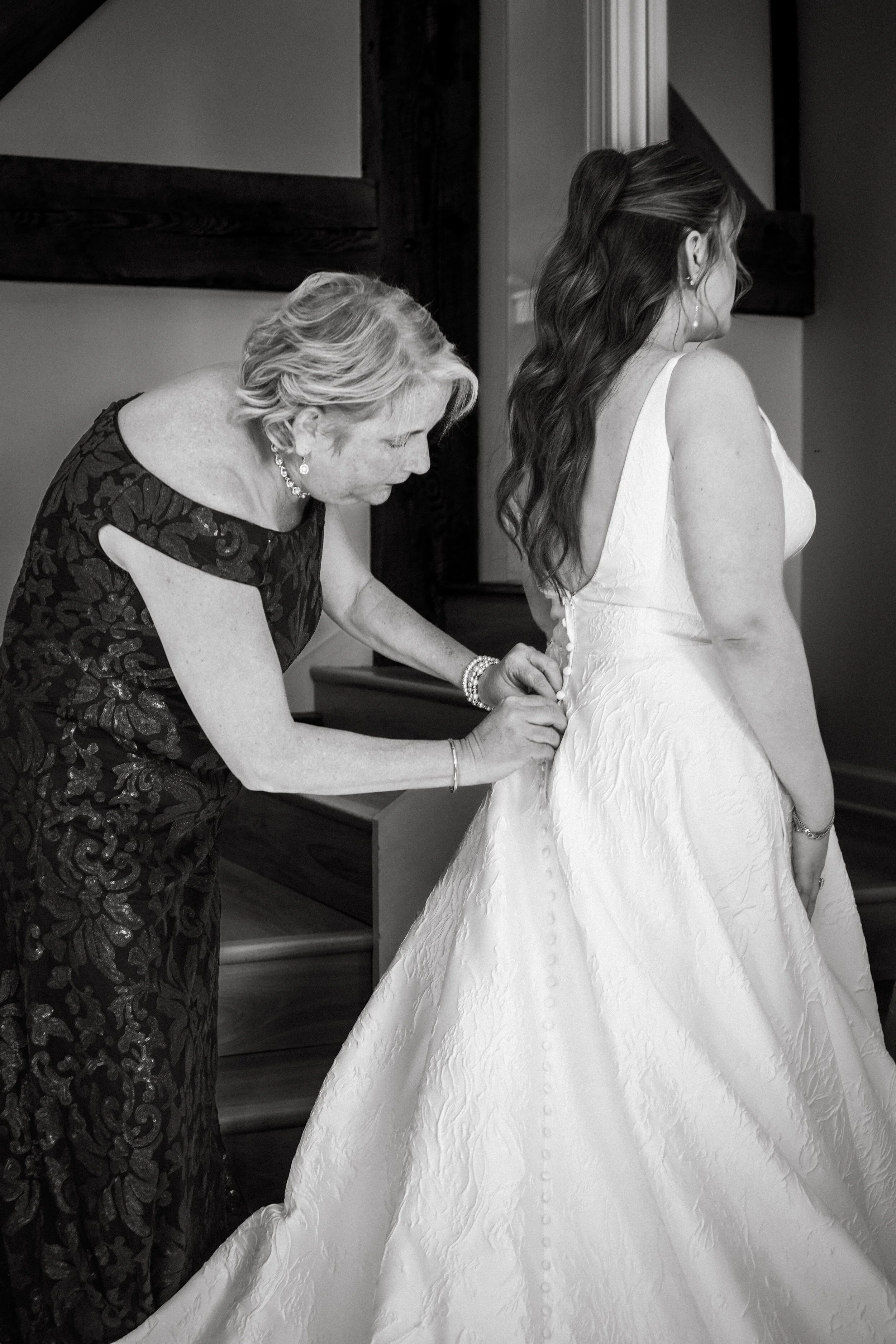 An elderly woman in a dark floral dress buttons a wedding gown on a young bride in a strapless dress.