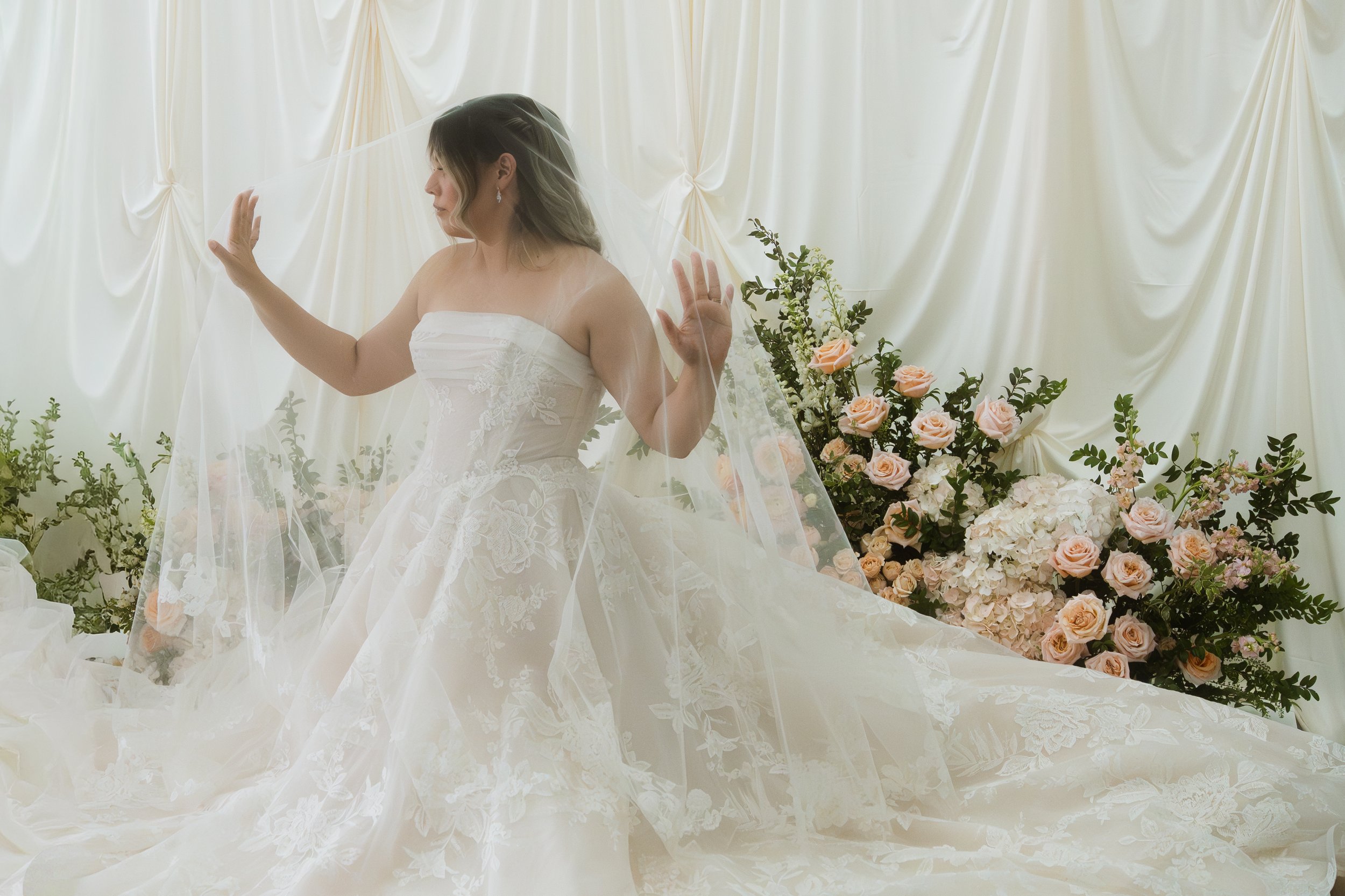 An older woman in a floral dress buttons up the back of a bridal gown worn by a young woman with long, wavy hair in a white sleeveless dress.