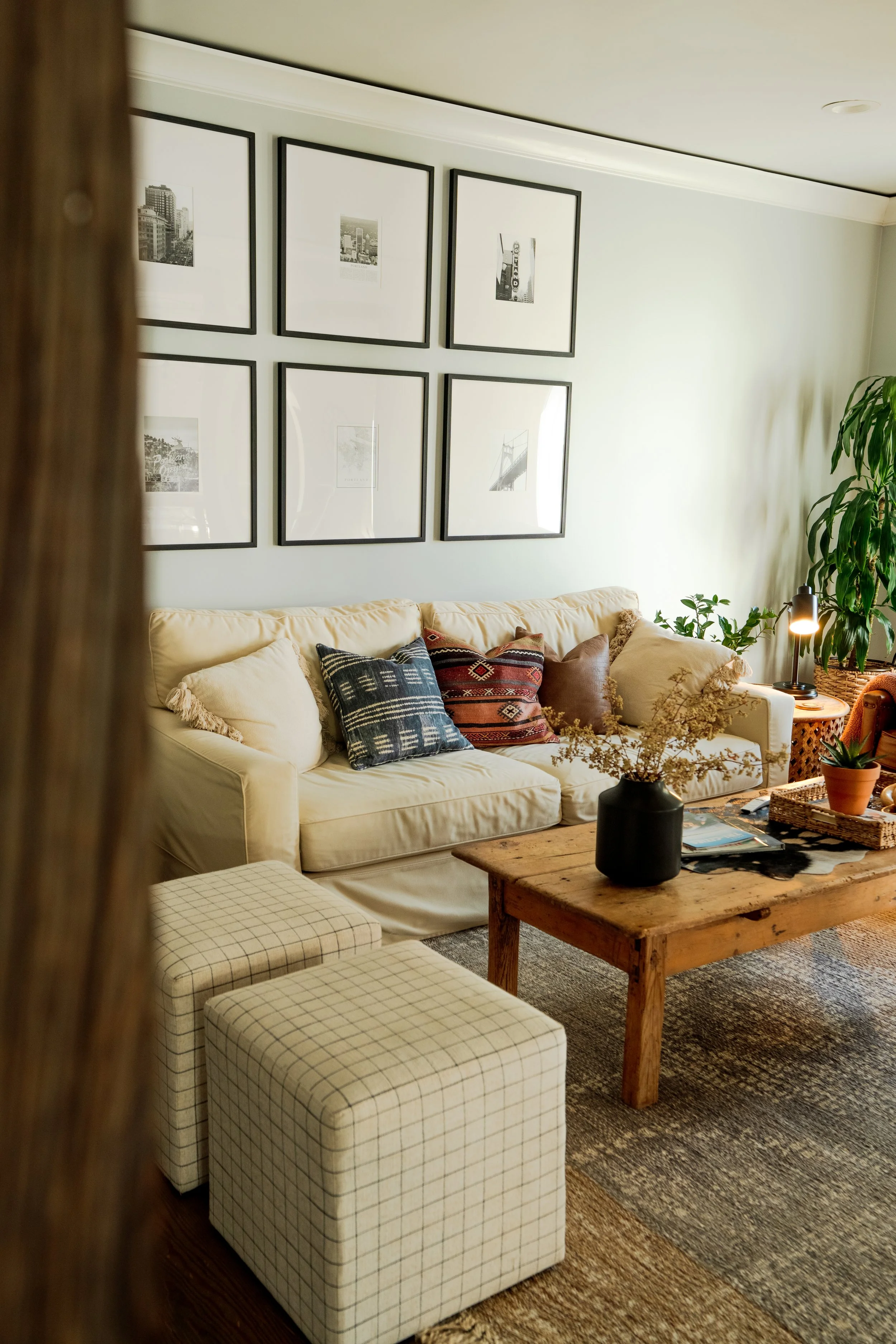 Cozy living room with cream sofa, decorative pillows, wooden coffee table with vase and plants, framed pictures on the wall, large green plant in corner, plaid ottomans, patterned area rug, and warm lighting.