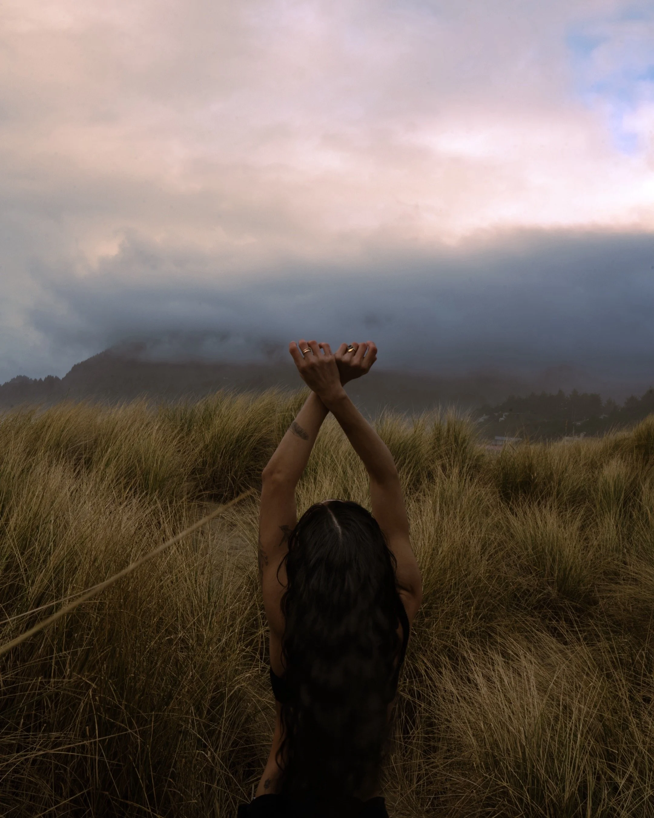 A person with long dark hair, seen from behind, standing in a grassy field with hills and cloudy sky in the background, raising their arms above their head.