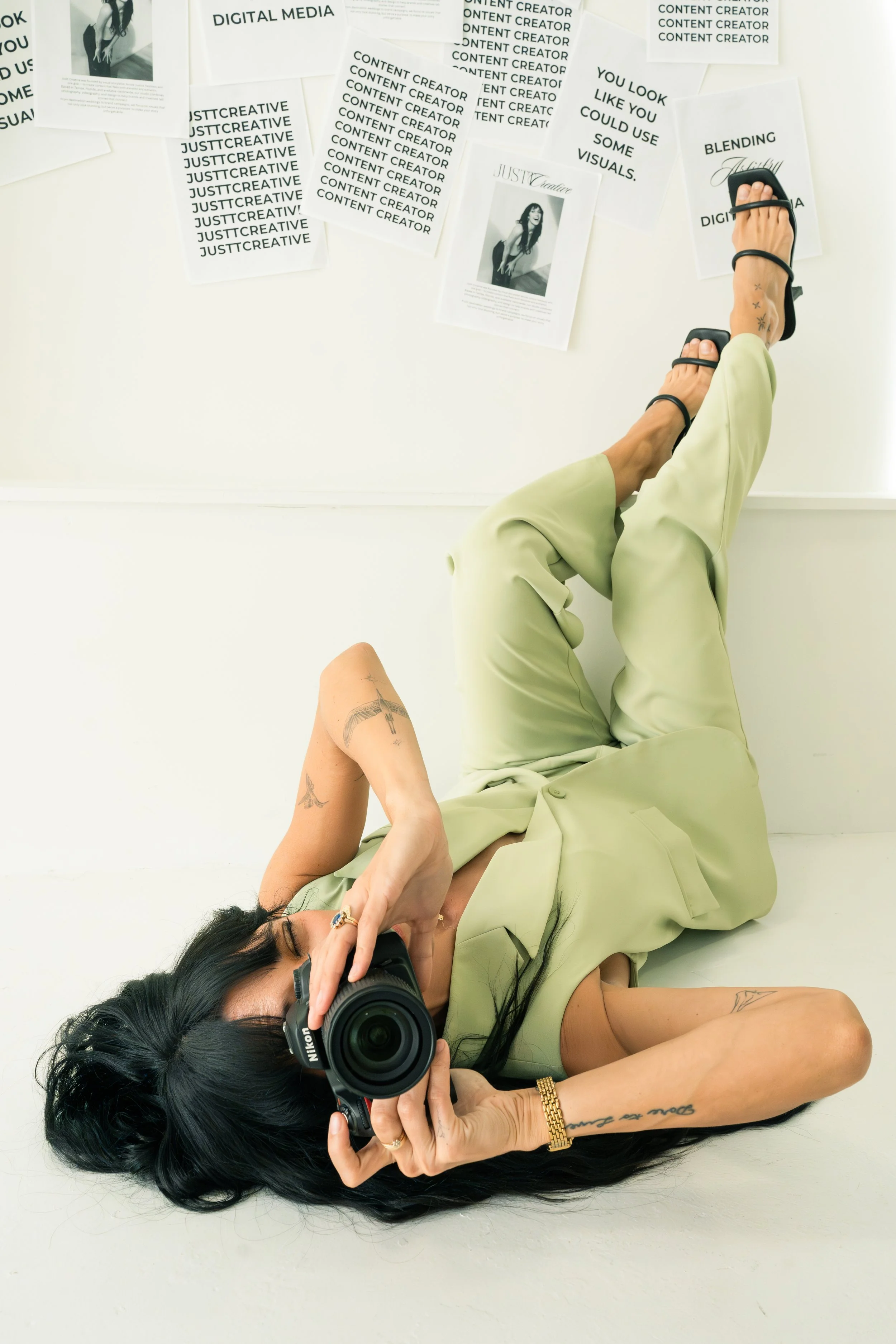 Woman lying on the floor taking a selfie with a camera, tattoos on her arms and legs, wearing a light green outfit and black sandals, with photos and posters on the wall behind her.