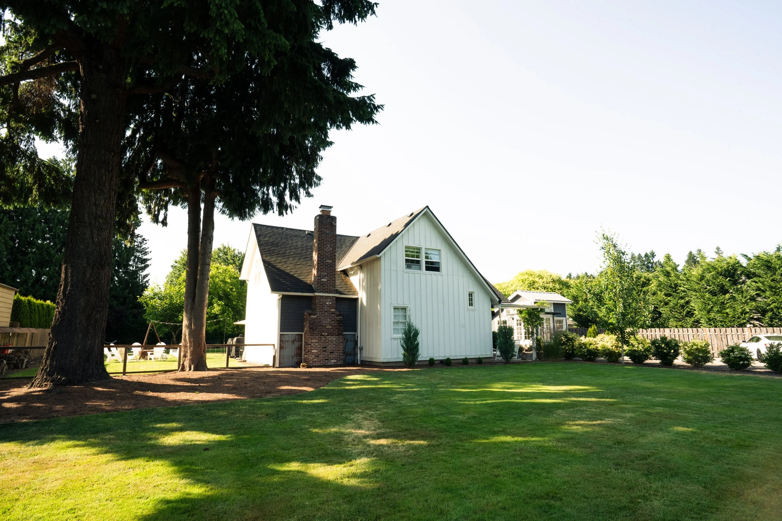 A house with a white wooden exterior and a chimney with a fenced backyard, surrounded by trees and a well-maintained lawn.
