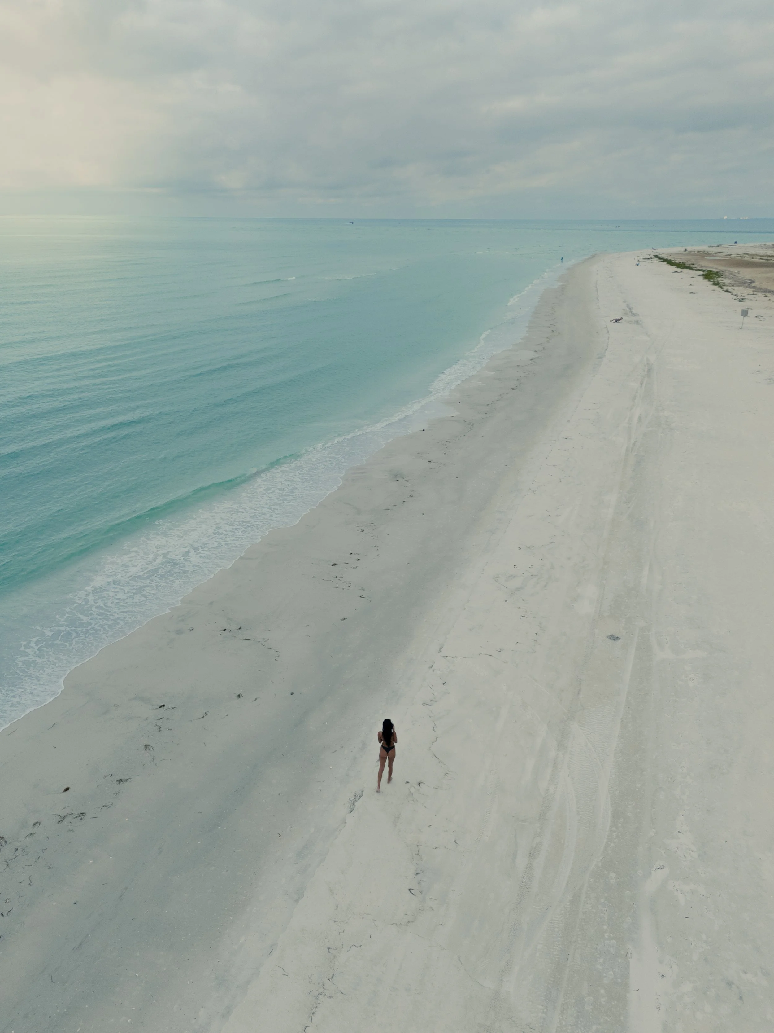 A woman in a swimsuit walking alone on a beach with white sand, near calm turquoise ocean waters, under an overcast sky.