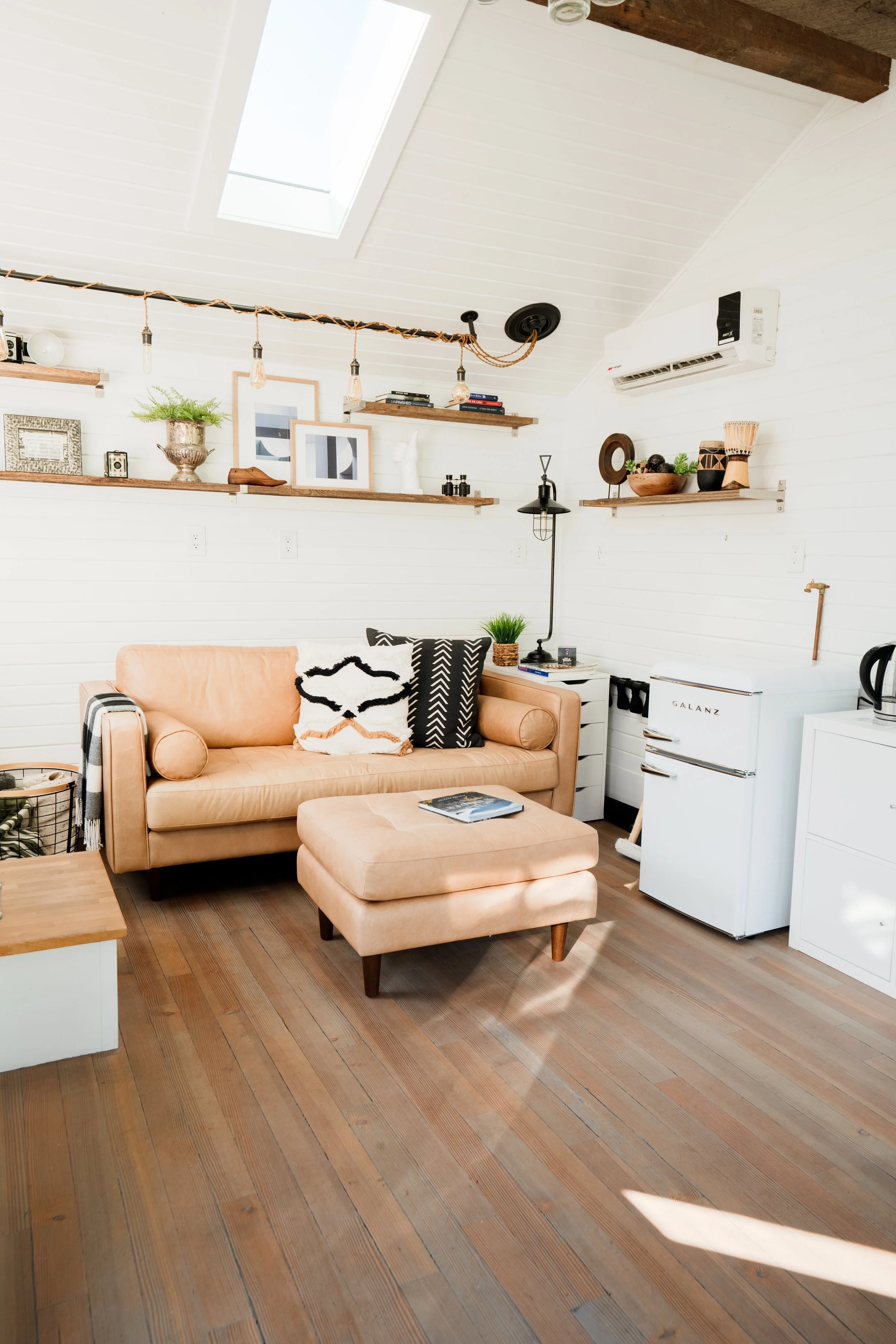 Bright living room with white paneled walls, a skylight, and wooden flooring. Features a peach-colored sofa with patterned pillows, a matching ottoman, and decorative shelves with artwork and plants.