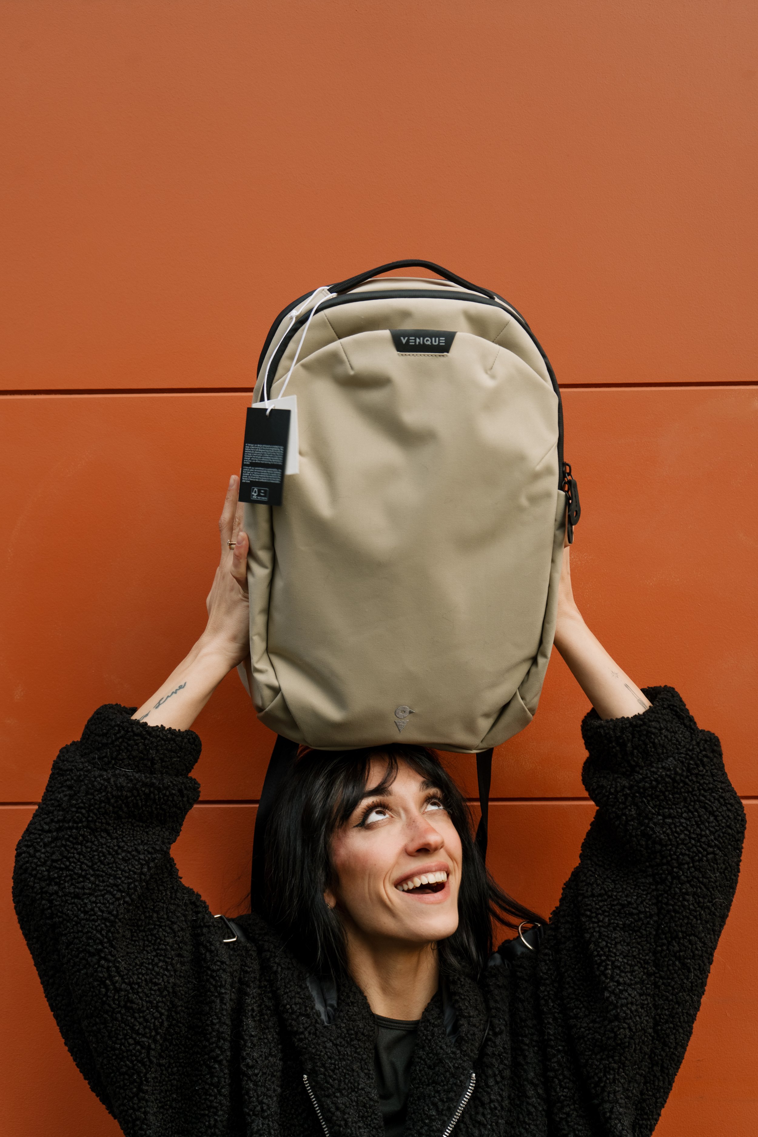 A woman with dark hair, smiling and looking up, holding a beige backpack above her head, standing in front of an orange wall.