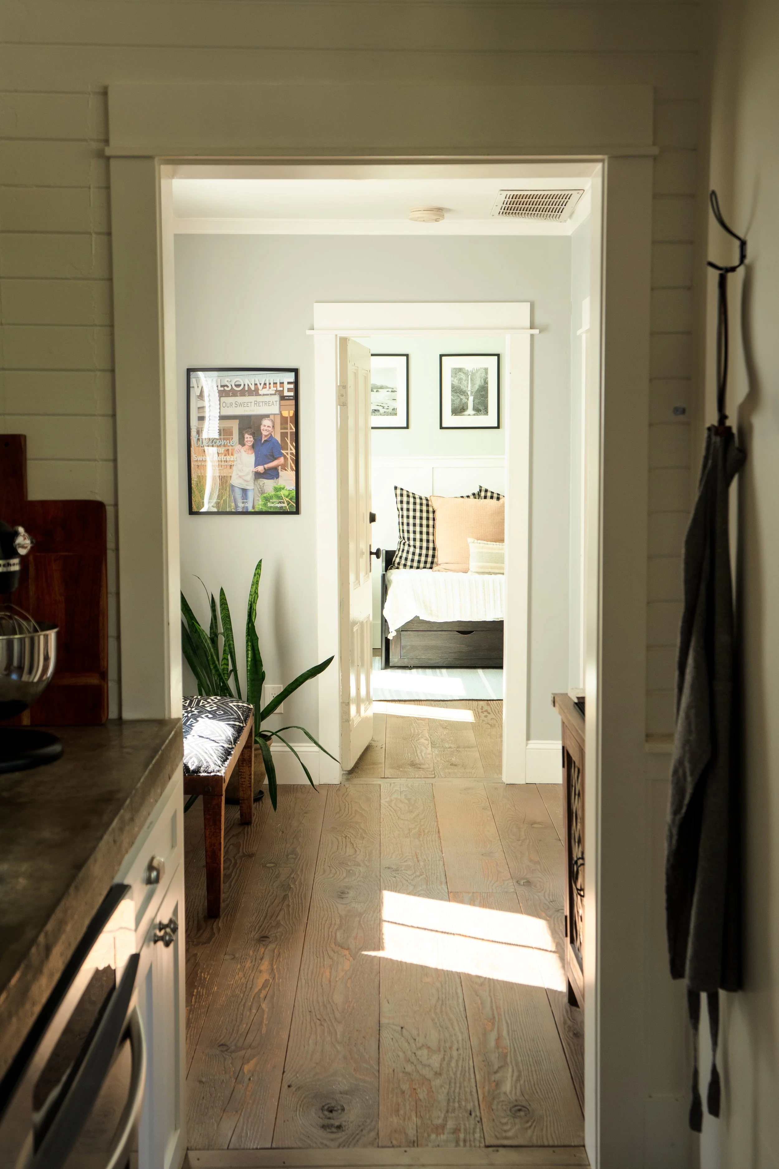 View through a doorway into a bright bedroom with framed photos on the wall and a bed with pillows, seen from a kitchen with wooden flooring and a counter.