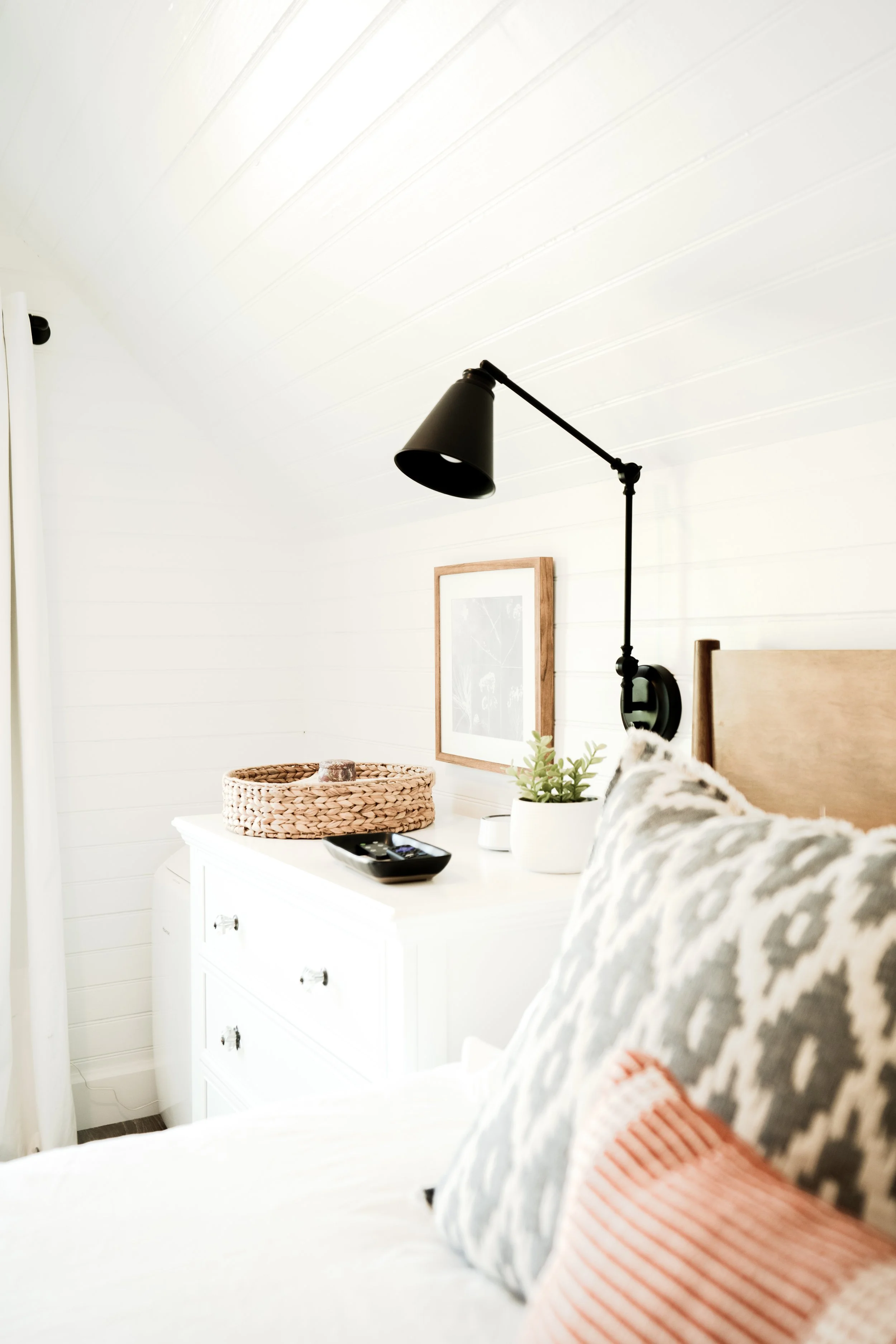 A cozy bedroom corner with a white dresser, decorative pillows, a wall-mounted black reading lamp, and a framed picture on a white paneled wall.