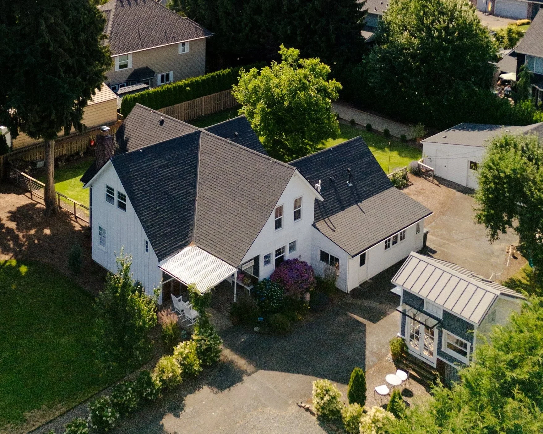 Aerial view of a two-story white house with a gabled roof, front porch, surrounded by trees, shrubs, and neighboring houses, with a backyard and driveway.