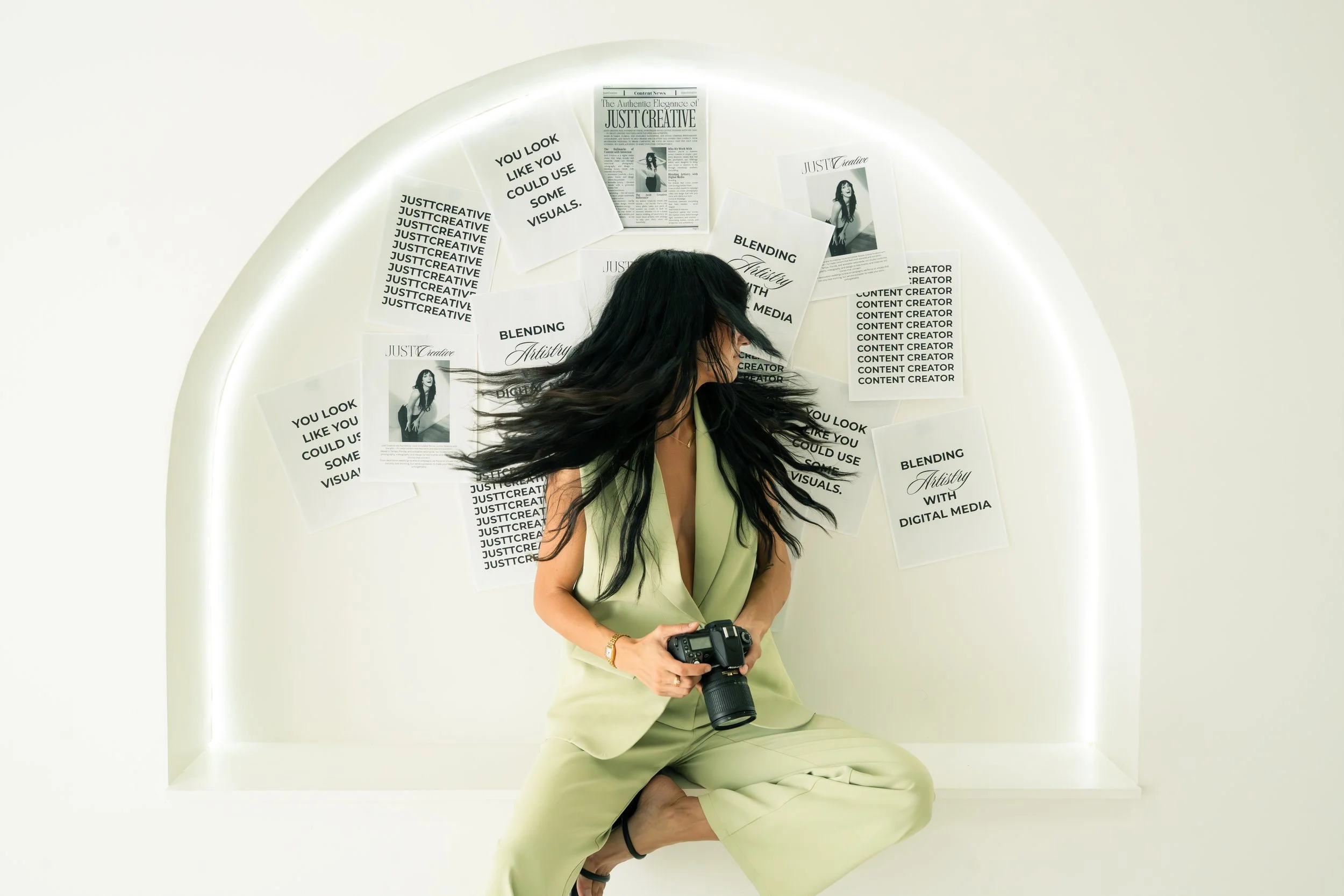 Woman with long black hair, dressed in a light green sleeveless blazer and matching pants, sitting cross-legged on the floor while holding a camera. She is in front of a white wall with various black-and-white posters and printed pages, some of which have text like "You look like you could use some visuals" and "Blending artistry with digital media." The posters are arranged in a semi-circular pattern behind her.
