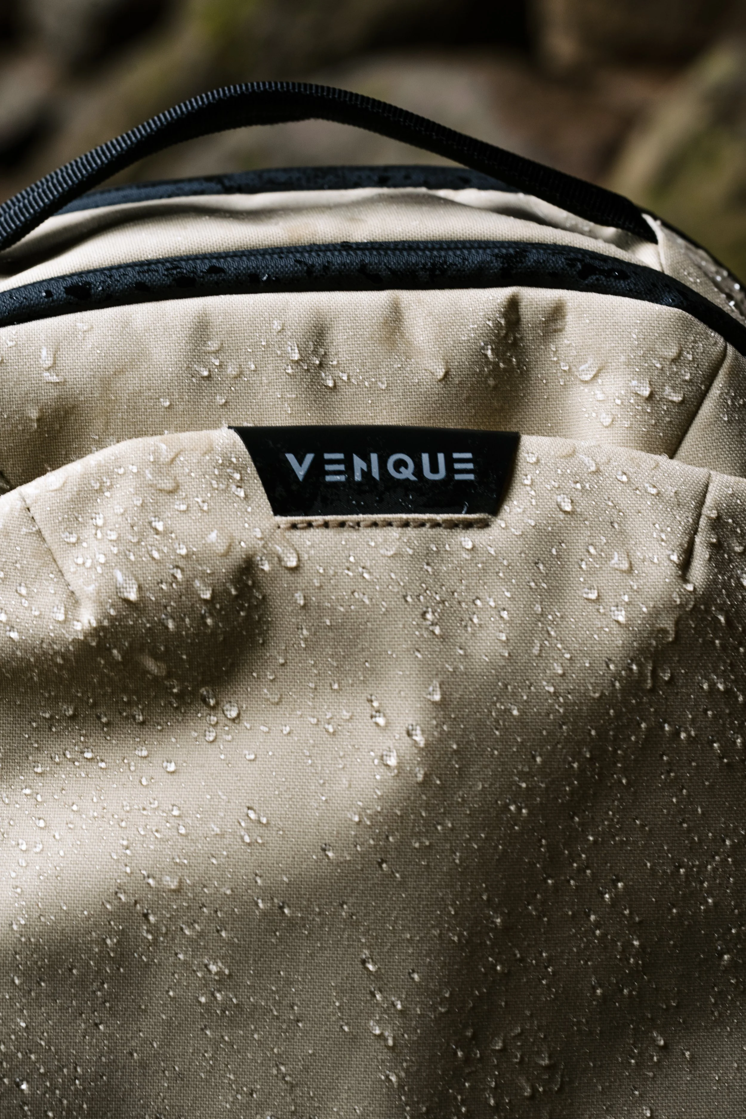 Close-up of a beige waterproof backpack with water droplets on its surface, displaying the brand name 'Venque' on a black label.
